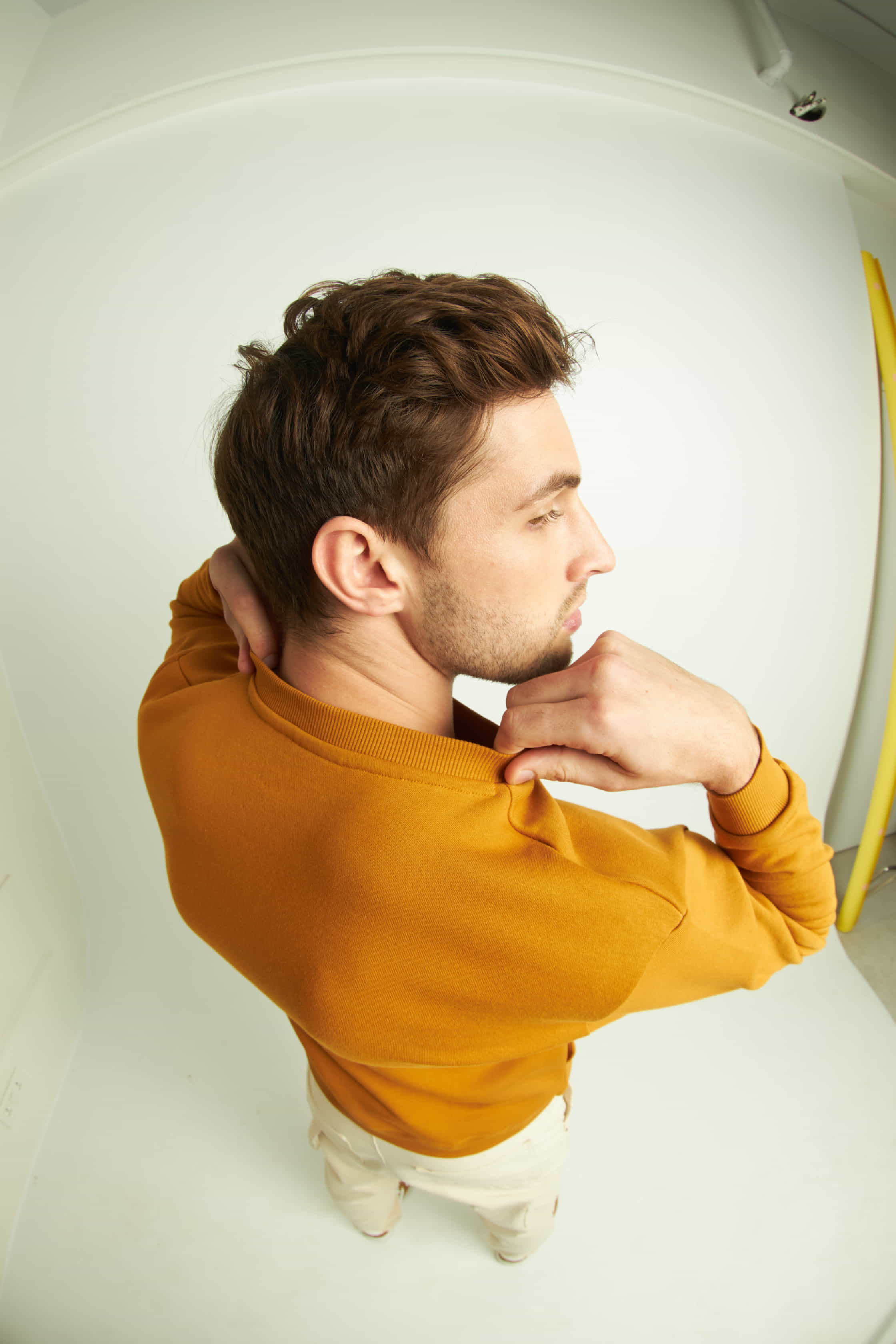 Man in a mustard yellow sweater standing against a white wall.