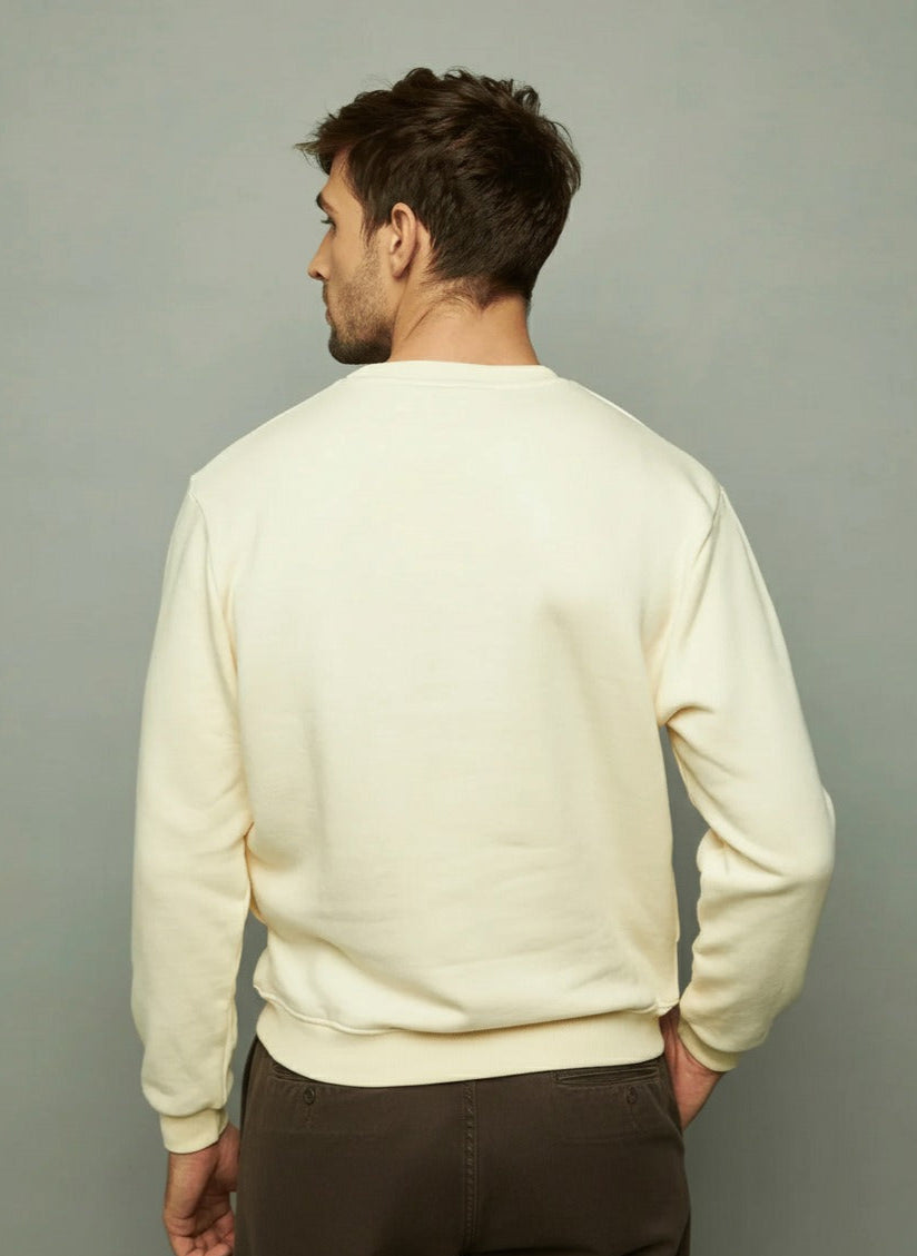 Man wearing an ecru sweatshirt and brown pants holding a brown jacket against a gray background